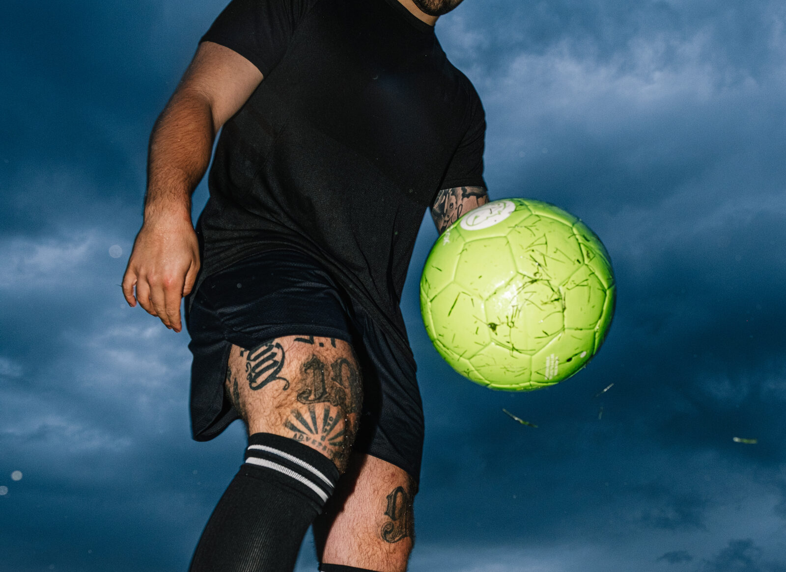 Fussballspieler jongliert mit einem gruenen Ball vor blauem Abendhimmel